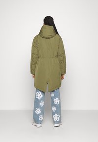 Persona con largas trenzas que lleva un abrigo verde oliva con capucha y jeans azules con grandes estampados de flores blancas, de pie ante un fondo sencillo.