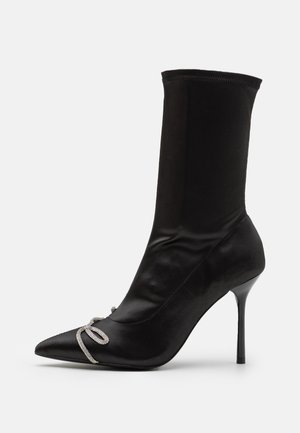 Bottine noire pointue à talon stiletto avec tige en tissu ajustée et nœud décoratif en strass argentés sur le bout.