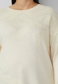 Close-up van een crèmekleurige gebreide trui met gestructureerde bloemmotieven, gedragen door een persoon, met zicht op de halslijn en bovenlichaam.