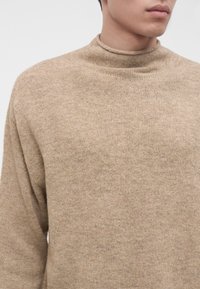 Jeune personne portant un pull en maille beige à col montant, montrant le haut du torse et une partie du visage sur un fond uni.