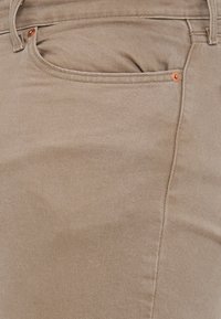 Pantalons en coton beige avec une texture lisse, présentant un tissage sergé diagonal, une poche avant et des accents en rivets cuivrés.