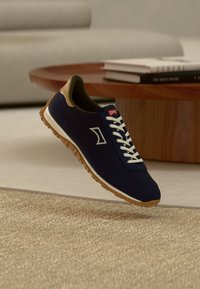 Αθλητικό παπούτσι σε ναυτικό μπλε με λευκά κορδόνια, επάνω μέρος από υφασμάτινο υλικό με υφή και καστανές λεπτομέρειες. Διαθέτει ελαφριά καφέ σόλα από καουτσούκ και λογότυπο στο πλάι.