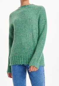 Suéter de punto verde con cuello redondo, con puños y dobladillo acanalados, usado con jeans azules por una persona contra un fondo blanco.