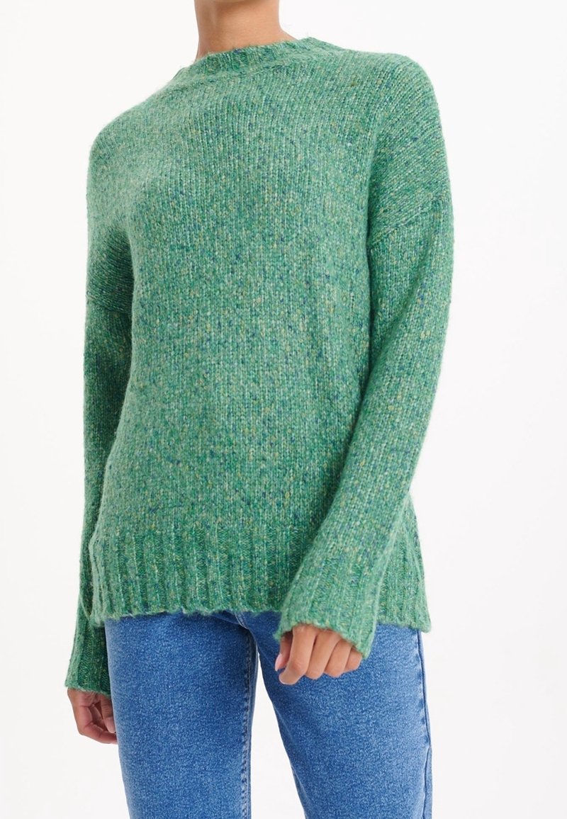 Suéter de punto verde con cuello redondo, con puños y dobladillo acanalados, usado con jeans azules por una persona contra un fondo blanco.