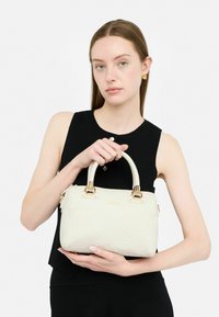 Piccolo borsa a tracolla in crema con logo impresso, accessori in oro, design strutturato e doppio manico superiore, tenuta da una persona che indossa un outfit nero.