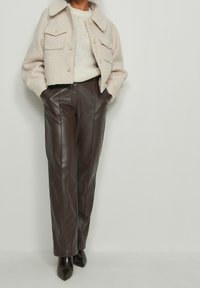 Osoba ubrana w kremową wełnianą kurtkę, kremowy sweter z dzianiny, ciemnobrązowe skórzane spodnie i brązowe, spiczaste buty, stojąca na tle jednolitego tła.