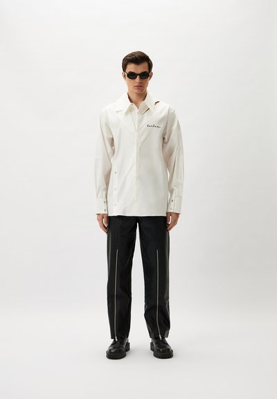 White long-sleeve shirt with a collar and black stitching; paired with black pants featuring zip details and chunky black shoes.