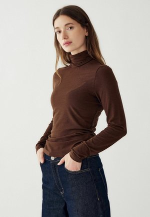 Tricou cu guler înalt de culoare maro, realizat dintr-un material moale și elastic, având mâneci lungi și o siluetă cusută, asortat cu blugi de culoare albastru închis.