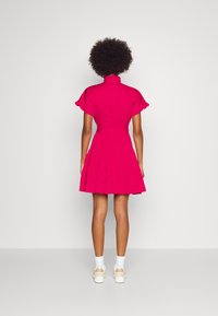 Kvinna med afrohår som bär en kortärmad, ljusrosa volangklänning, vita strumpor och beige sneakers, stående vänd bort.