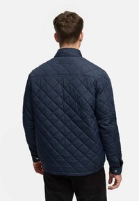 Dunkelblau gesteppte Jacke mit Stehkragen, ausgestattet mit Rautensteppung, Druckknöpfen und einem Logo auf dem Rücken. Glatte Textur.