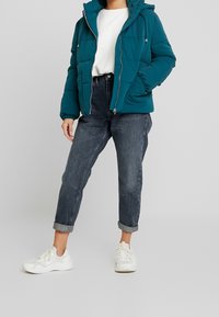 Vihreä puffer-takki hupulla, vetoketjulla edessä, kaksi taskua ja joustavat hihansuut. Yhdistetty ylös käännettyjen tummien farkkujen ja valkoisten tennareiden kanssa.