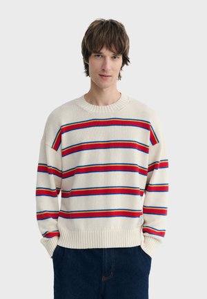 Young man with brown hair wearing a cream sweater with red and blue horizontal stripes, hands in dark blue pants pockets.