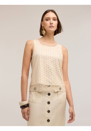 Frau trägt ein ärmelloses beiges Oberteil mit kleinen Verzierungen und einen beigen Rock mit Knöpfen, kombiniert mit auffälligen Ohrringen und Armbändern.