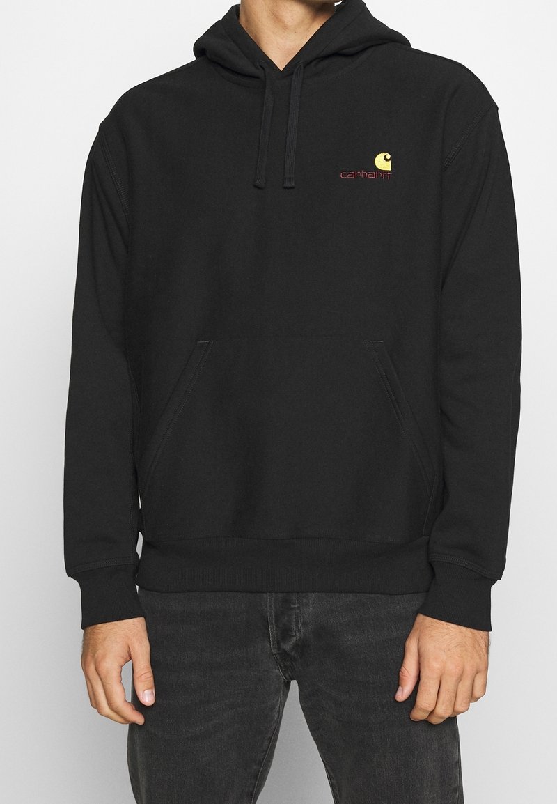 Sweat à capuche noir avec poche kangourou à l'avant et petit logo Carhartt jaune sur le côté gauche de la poitrine, porté avec un jean foncé à partir de la taille.