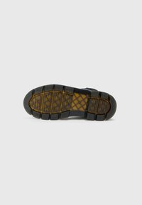 Semelle de chaussure en caoutchouc noir avec une surface texturée, présentant un motif de nid d'abeille et des crampons épais et surélevés pour une meilleure adhérence. Détails en marron.