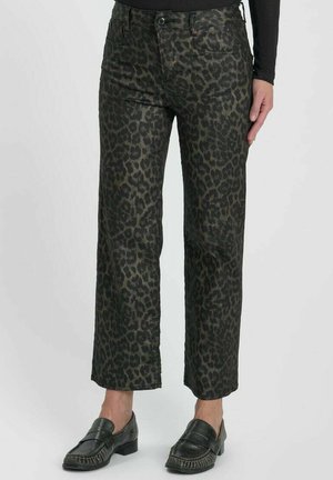Vrouw draagt cropped donkergroene en zwarte luipaardprint broek met zwarte slip-on loafers, staand tegen een effen achtergrond.