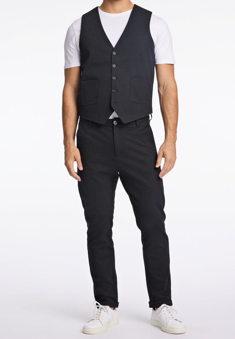 Homme portant un gilet noir ajusté et un pantalon sur un t-shirt blanc à manches courtes avec des baskets blanches, debout sur un fond blanc.
