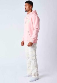 Felpa rosa con tasca frontale e logo a forma di cuore, abbinata a pantaloni in denim bianco strappati con strappi orizzontali. Sneakers bianche completano l'outfit.