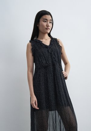 Ung kvinde i en ærmeløs sort kjole med hvide polkaprikker og flæseærmer, stående mod en lysegrå baggrund.