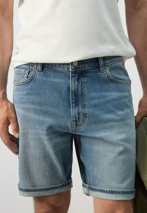 Mand iført lyseblå denimshorts med opsmøgede kanter og en hvid skjorte, der holder en olivengrøn jakke i højre hånd.