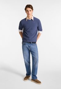 Navy ribbeltrui met een crèmekleurig overhemd eronder, gecombineerd met lichtblauwe rechttoe-rechtaan jeans en bruine suède instappers.