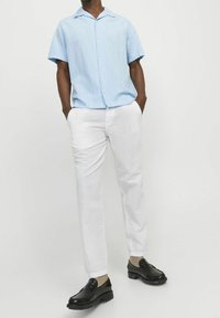 Man som bär en ljusblå kortärmad skjorta, vita byxor, beige strumpor och svarta loafers står med händerna i fickorna mot en enkel bakgrund.