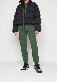 Svart puffjacka med en kortad design, tillsammans med gröna sweatpants med en logo. Svart och grå höga sneakers fulländar outfiten.