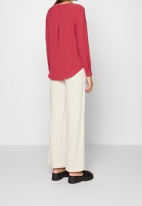 Camicia da donna a maniche lunghe di un rosso intenso, con una piega sul retro, orlo curvato e polsini con bottone, abbinata a pantaloni bianchi larghi e sandali neri.
