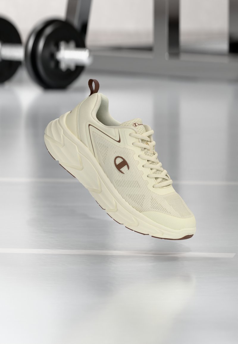 Αθλητικό παπούτσι σε μπεζ απόχρωση με παχιά σόλα και καφέ λογότυπο αιωρείται πάνω από το δάπεδο του γυμναστηρίου με αλτήρες θολωμένους στο φόντο.