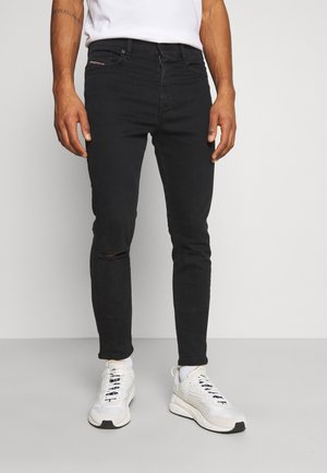 Homme portant un jean slim noir avec une déchirure au genou droit, des chaussettes blanches, des baskets blanches et un t-shirt blanc rentré dans le pantalon.