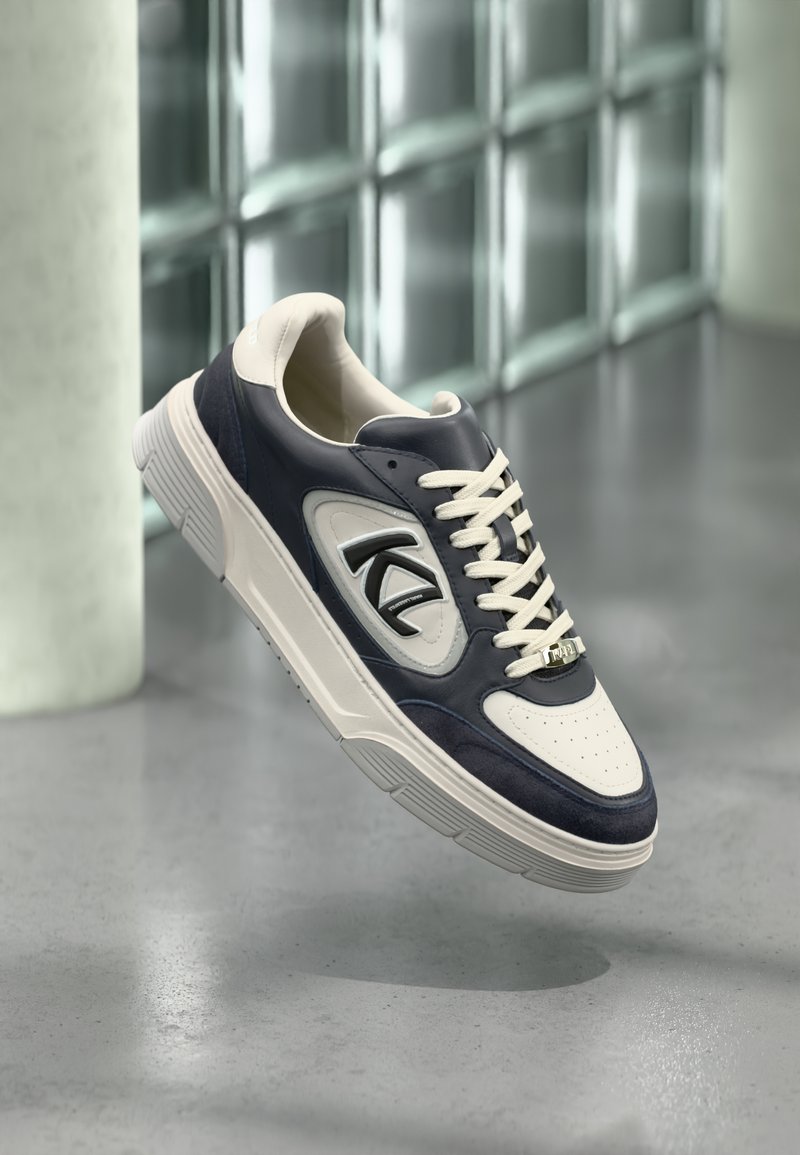 Αθλητικό παπούτσι σε ναυτικό μπλε και κρεμ χρώμα με δερμάτινα και σουέτ υλικά, διάτρητη μύτη, λαστιχένια σόλα και εμφανές λογότυπο στο πλάι.