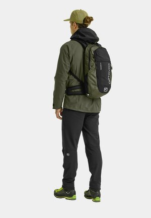 Black and green backpack with textured finish, worn by a person in a green jacket and black pants, standing against a plain background.