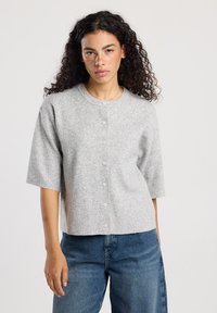 Woman with curly hair wears light gray buttoned short-sleeve cardigan and blue jeans, standing against plain light background.