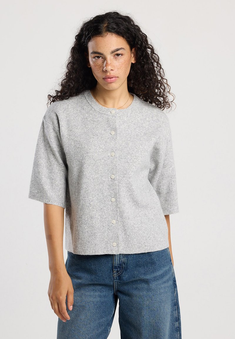 Woman with curly hair wears light gray buttoned short-sleeve cardigan and blue jeans, standing against plain light background.