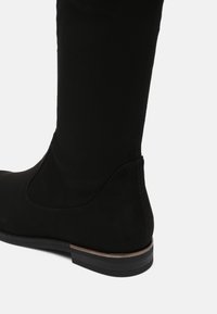 Botte noire montante jusqu'au genou avec talon bas empilé, texture lisse et détail de couture subtil, présentée sur un fond gris clair.