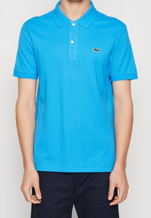 Homme portant un polo bleu vif à manches courtes avec deux boutons blancs et un petit logo crocodile vert sur la poitrine.