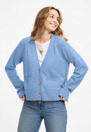 Young woman smiling, wearing a light blue cardigan over a white shirt and blue jeans, standing with hands on hips against a plain background.