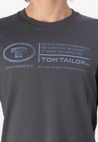 Camiseta de algodón gris con cuello redondo, que presenta texto y gráficos en azul, incluido el nombre de la marca "TOM TAILOR" y coordenadas impresas en la parte frontal.