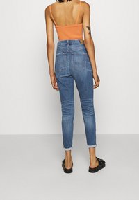 Högmidjade blå denimjeans med smal passform, uppvikta ärmar och bakfickor. Modellen bär en figurnära orange topp och svarta sandaler.