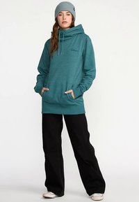 Sweat à capuche sarcelle avec col bénitier, cordons de serrage et poches latérales, associé à un pantalon large noir et un bonnet en tricot gris clair.