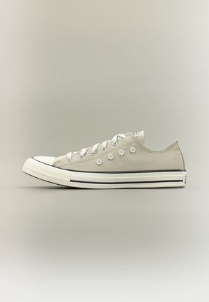 Sneaker beige in tela a taglio basso con suola in gomma bianca, lacci bianchi e sei piccole decorazioni a fiore bianco sul lato.