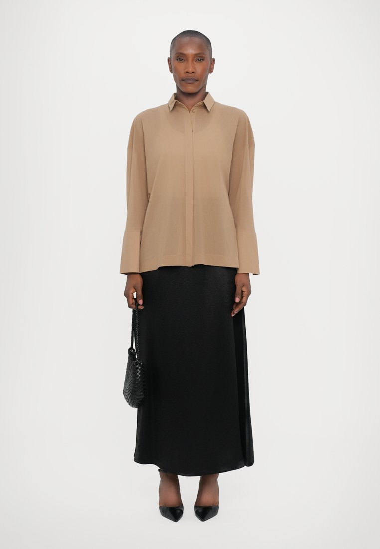 Chemise beige à manches longues avec col, associée à une jupe longue noire en satin. Comprend un sac à main noir texturé et des talons noirs pointus.