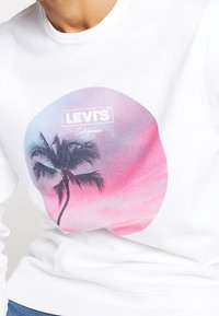 Sweat-shirt blanc avec un motif circulaire d'un palmier et un coucher de soleil dégradé en rose et violet, avec le logo "LEVI'S" au-dessus.