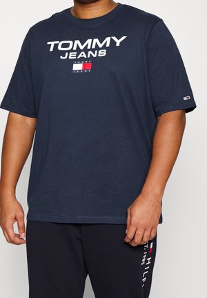 Persona que lleva una camiseta Tommy Jeans azul marino con logo blanco y pantalones oscuros a juego con marca lateral.