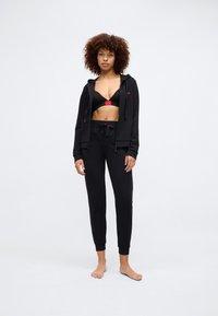 Black zip-up hoodie with red accents, paired with a black bra and black jogger pants. Smooth fabric, slim fit, ankle cuffs, and drawstring.