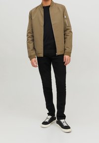 Khaki bomber jacket z zamkiem z przodu oraz boczną kieszenią, w komplecie z czarnym swetrem, czarnymi dżinsami i czarnymi trampkami z białymi sznurówkami.