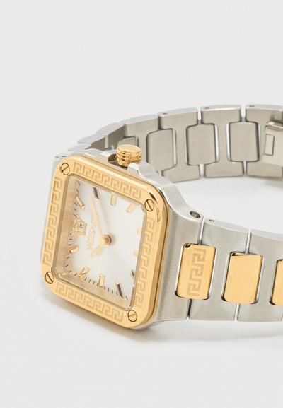 Kvadratinis sidabro ir aukso spalvos riešo laikrodis su baltu ciferblatu, aukso valandų žymekliais ir rodyklėmis, graikiškos rakto formos raštu ant apyrankės krašto ir grandinėlės dalių.