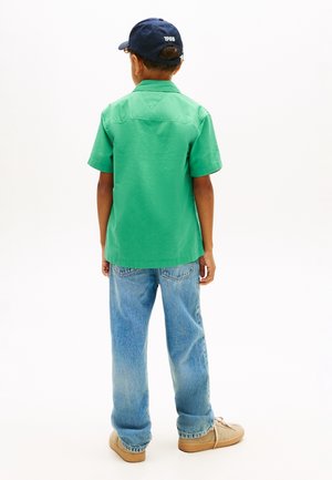 Gutt iført en grønn kortermet skjorte, blå jeans, beige joggesko og en marineblå caps, stående med ryggen mot på en hvit bakgrunn.