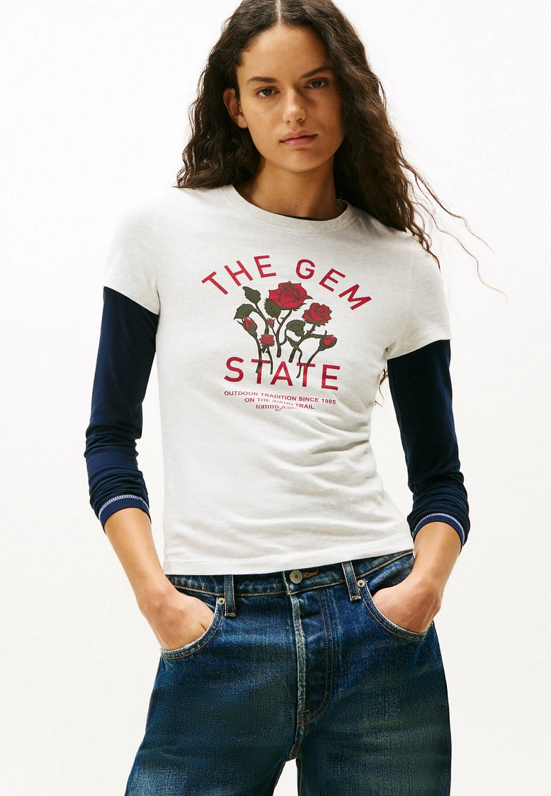 Camiseta gris claro con mangas cortas en navy, que presenta un gráfico de una rosa roja y el texto "EL ESTADO DE LAS GEMAS" en rojo. Combinada con jeans azules.