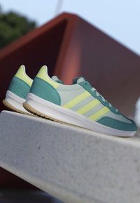 Paar von türkisfarbenen und hellgelben Sneakers mit weißen Sohlen, die auf einer Betonmauer im Freien vor verschwommenem geometrischem Hintergrund ruhen.